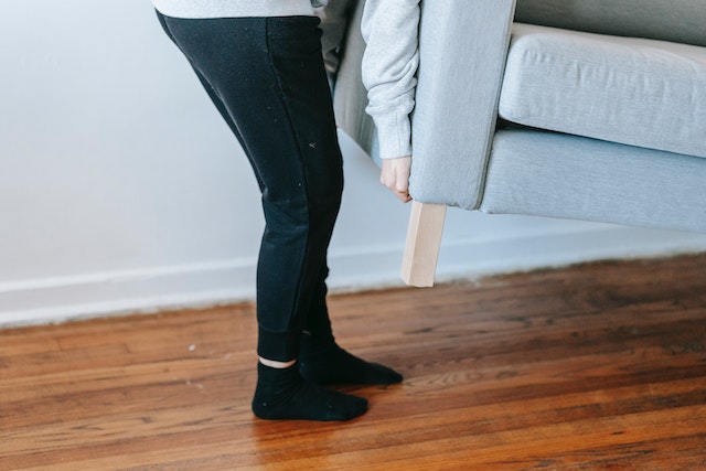 A tenant in black pants and socks lifts one side of a grey couch off a hardwood while moving out of their rental