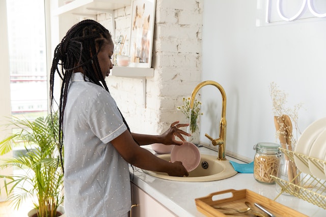 a tenant with box braids washes their dishes in a kitchen sink set into a white marble countertop with a gold faucet copy